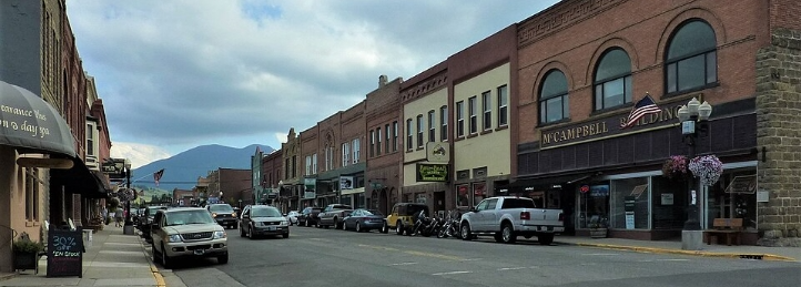 Historic downtown street with brick storefronts, parked vehicles, hanging flowers, and mountains beneath a cloudy sky.