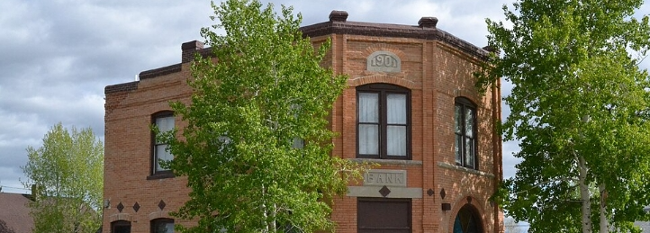 Historic 1901 red brick bank building framed by leafy green trees beneath a cloudy sky.